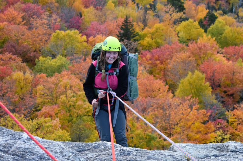 In the image, a woman wearing a helmet and a backpack is rappelling down a rock face. She is smiling and appears to be enjoying the activity. The background is filled with colorful autumn foliage, creating a vibrant and scenic setting. The composition of the image captures a moment of adventure and the beauty of nature.